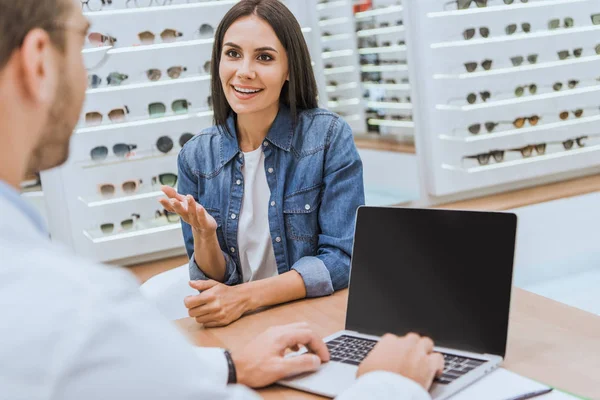男性验光师的部分看法在笔记本电脑上 而女人手指着眼科商店 — 图库照片