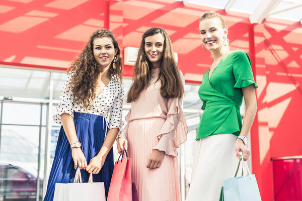 низкий угол обзора красивых стильных молодых женщин, держащих бумажные пакеты и улыбающихся на камеру в торговом центре
 