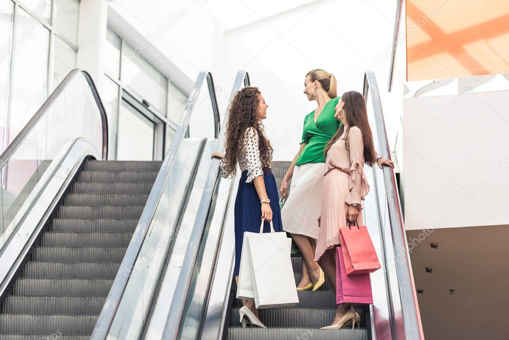 low angle view of beautiful smiling young women holding shopping bags on escalator 