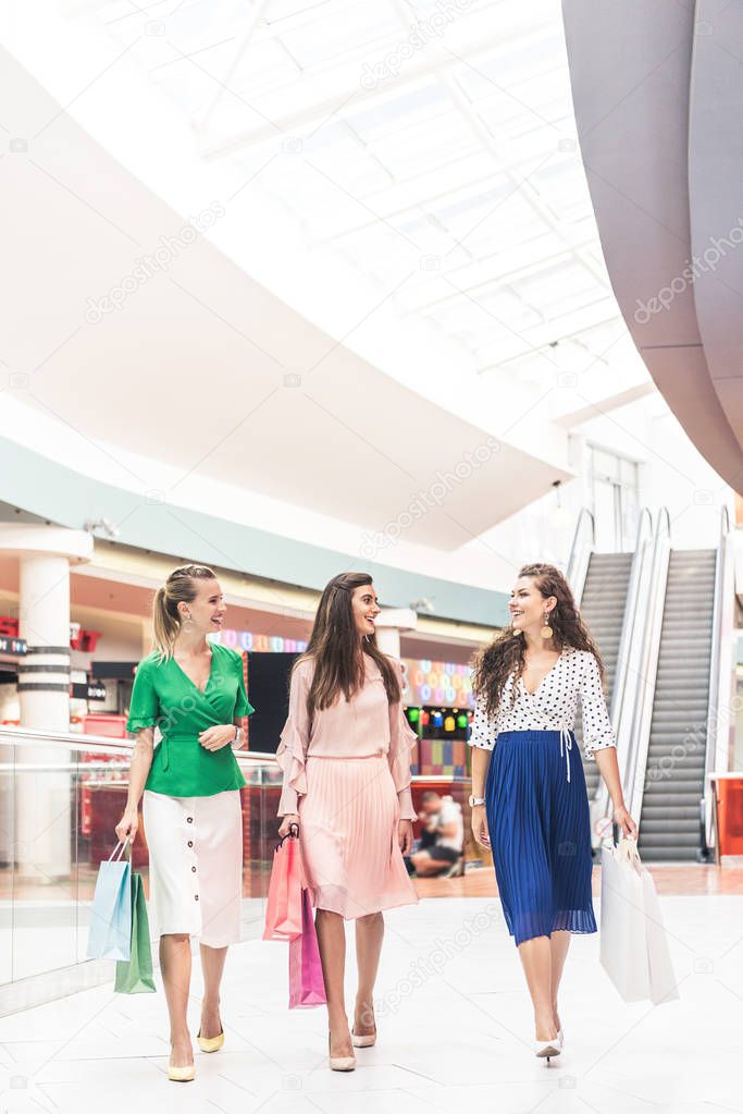 beautiful smiling young women holding paper bags and talking while walking together in shopping mall