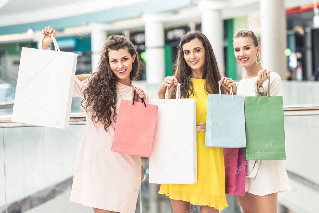 three beautiful happy girls holding shopping bags and smiling at camera in mall