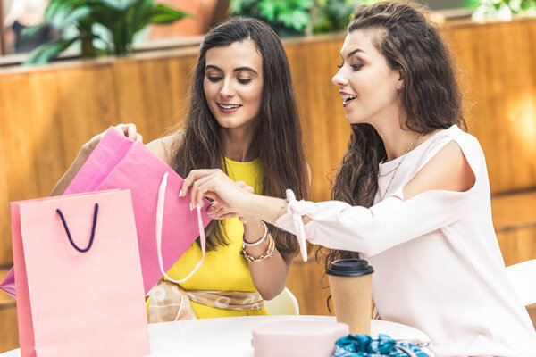 улыбающиеся молодые женщины, заглядывающие в сумку, сидя за столом в торговом центре
