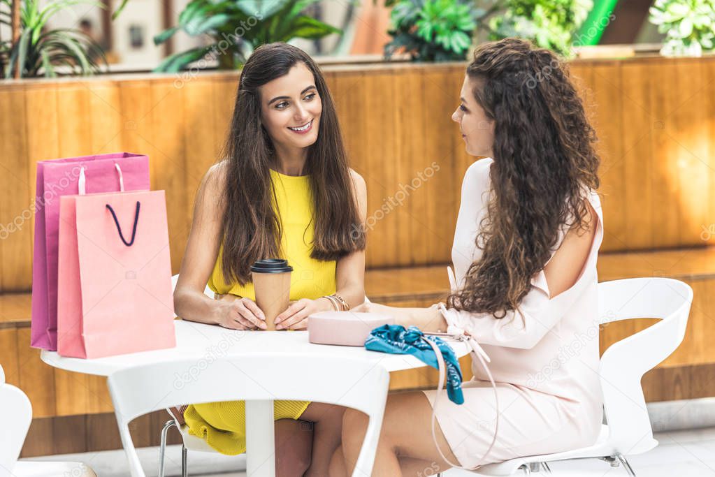 stylish young women smiling at talking while sitting in cafe at shopping mall