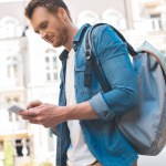 Вид снизу красивого молодого человека с рюкзаком, идущего по улице и использующего смартфон
