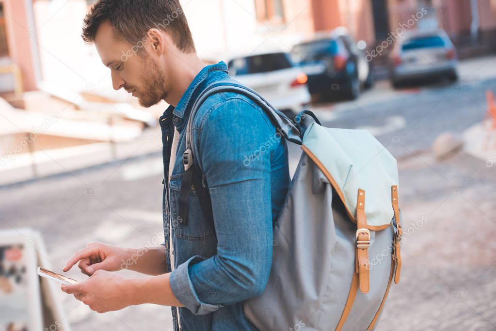 handsome young man with backpack walking by street and using smartphone