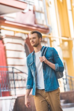 attractive young tourist with backpack walking by street and looking away clipart