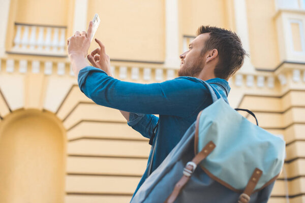 handsome young tourist with backpack taking photo with smartphone on street