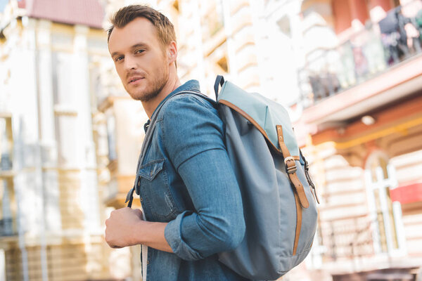 handsome young man with backpack walking by street and looking at camera