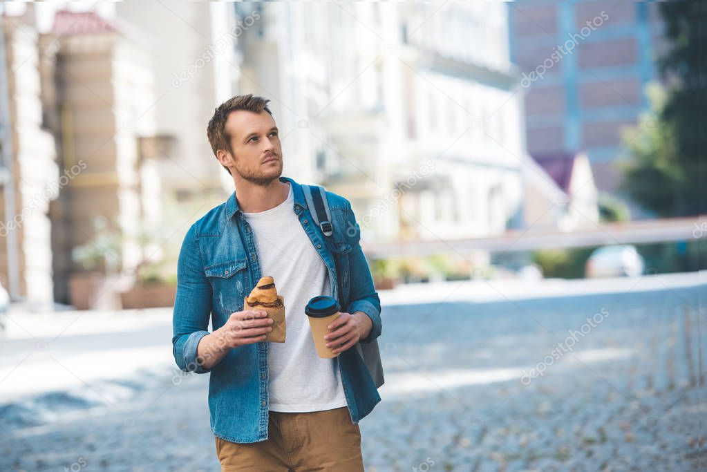 handsome young man with backpack, coffee to go and croissant walking by street and looking away