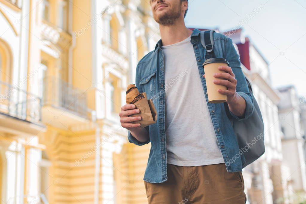 cropped shot of happy man with backpack, coffee to go and croissant walking by street