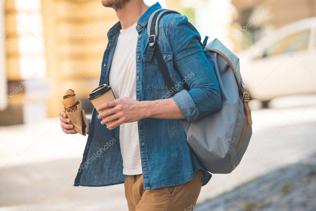 cropped shot of man with backpack, coffee to go and croissant walking by street