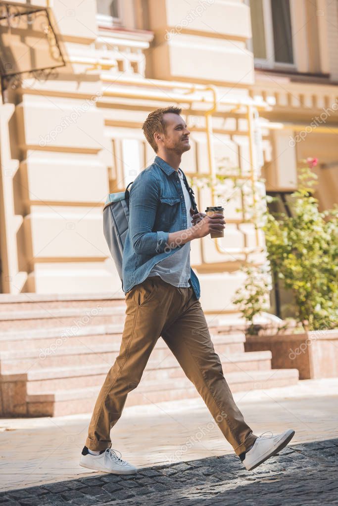 casual young man with coffee to go and backpack walking by street and looking away