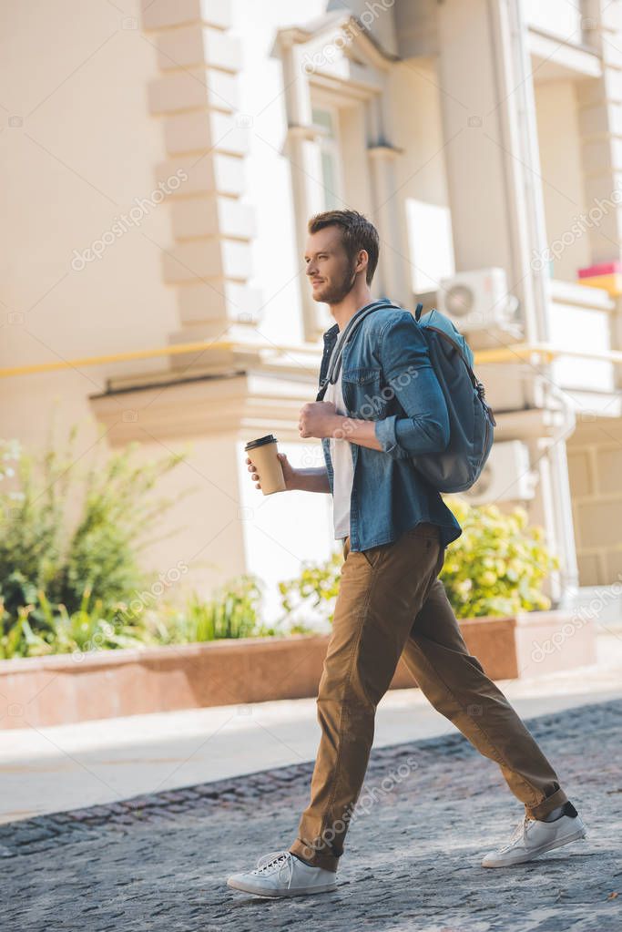 attractive young man with coffee to go and backpack walking by city