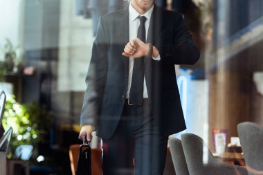 partial view of businessman with suitcase checking time while walking in cafe clipart