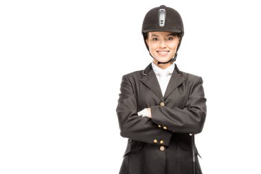 happy young horsewoman in uniform and helmet looking at camera with crossed arms isolated on white clipart
