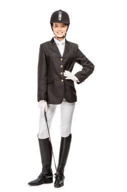 smiling young horsewoman in uniform holding horseman stick and looking at camera isolated on white clipart