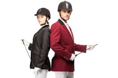 serious young equestrians in uniform and helmets holding horseman sticks while leaning back to back and looking at camera isolated on white  clipart