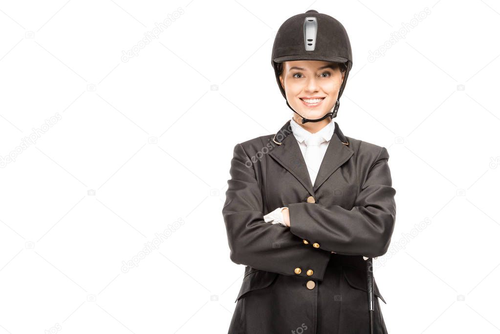 happy young horsewoman in uniform and helmet looking at camera with crossed arms isolated on white