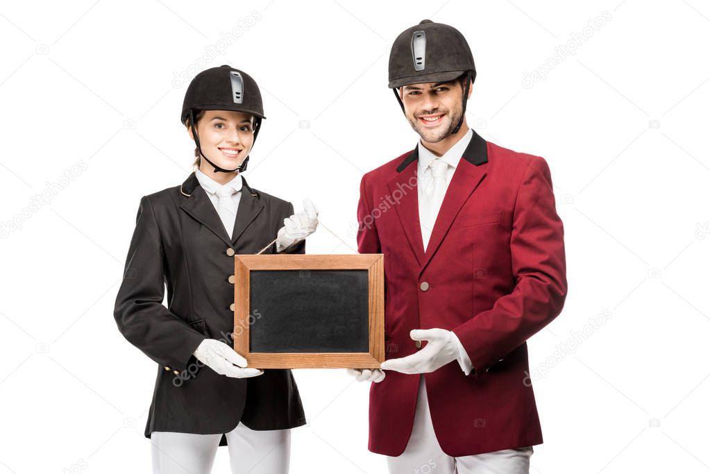 smiling young equestrians in uniform and helmets holding blank board and looking at camera isolated on white