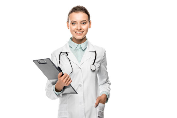 cheerful young female doctor in medical coat holding clipboard isolated on white