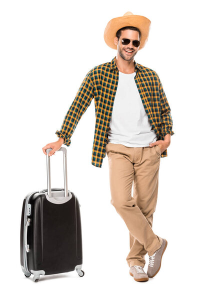 happy young male traveler in sunglasses and straw hat standing with wheeled bag isolated on white