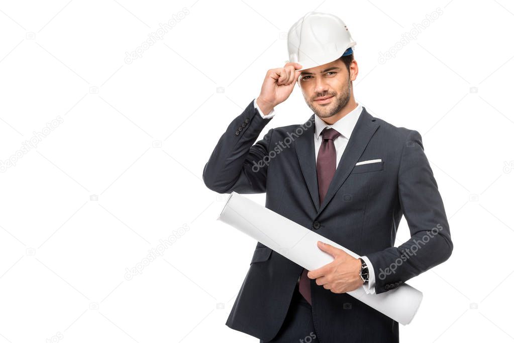 happy young male architect in suit holding blueprints and adjusting hard hat isolated on white 