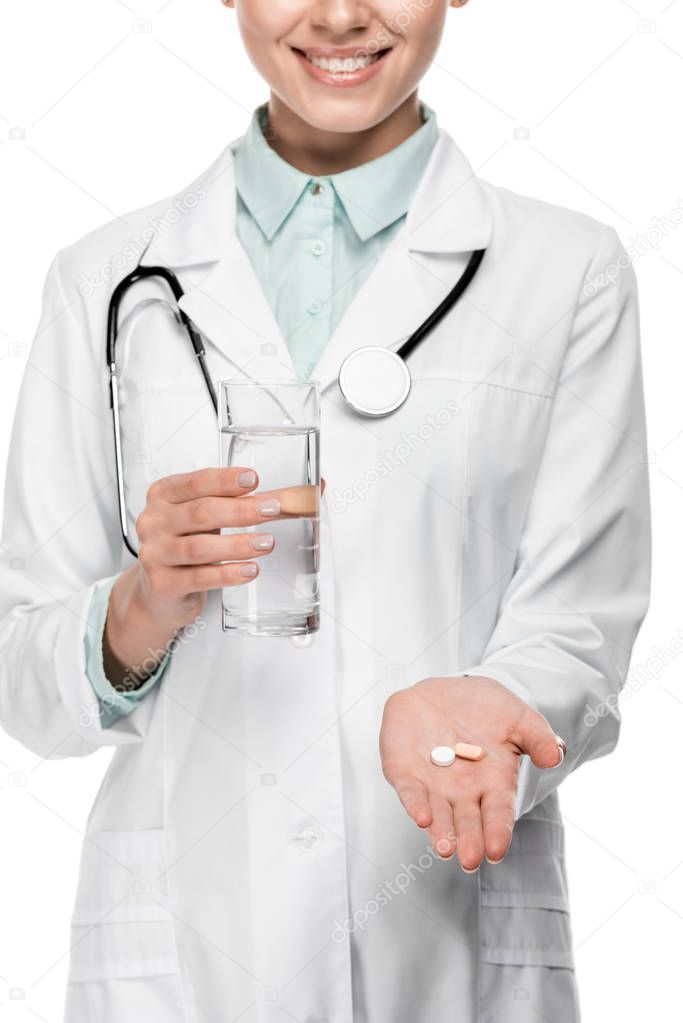 partial view of female doctor in medical coat holding glass of water and pills isolated on white