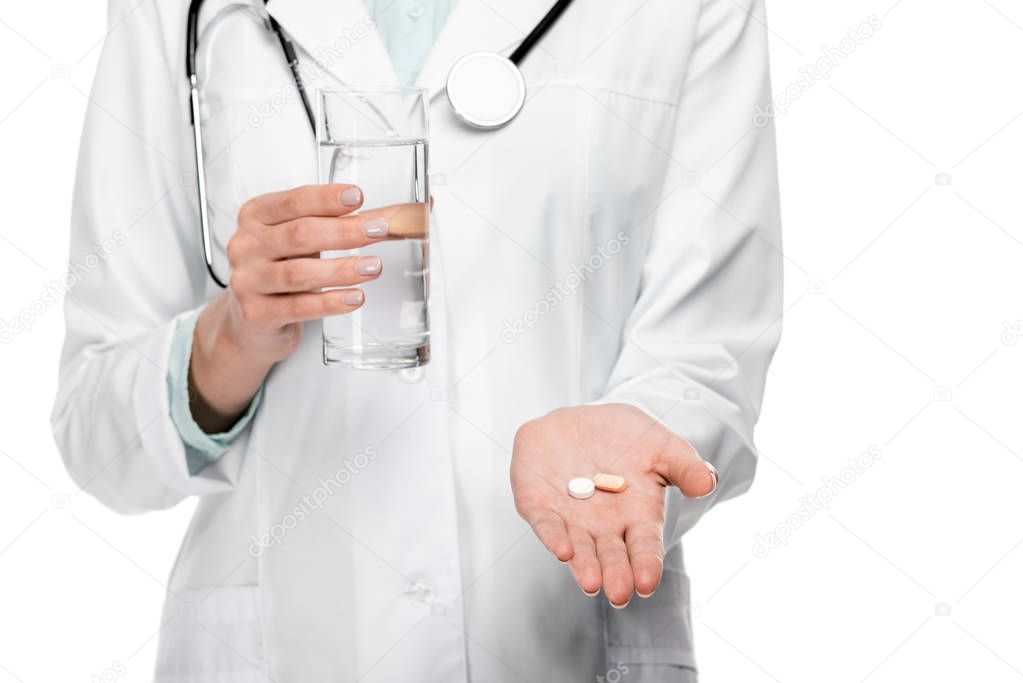 cropped image of female doctor in medical coat holding glass of water and pills isolated on white