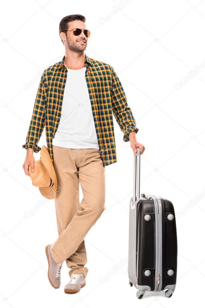 smiling young male traveler in sunglasses standing with wheeled bag isolated on white