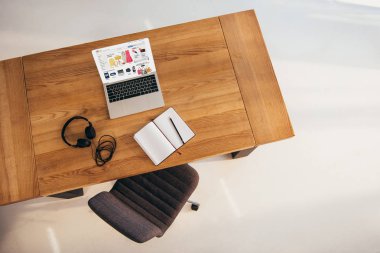 top view of laptop with ebay website, headphones and notebook on wooden table with office chair near by clipart