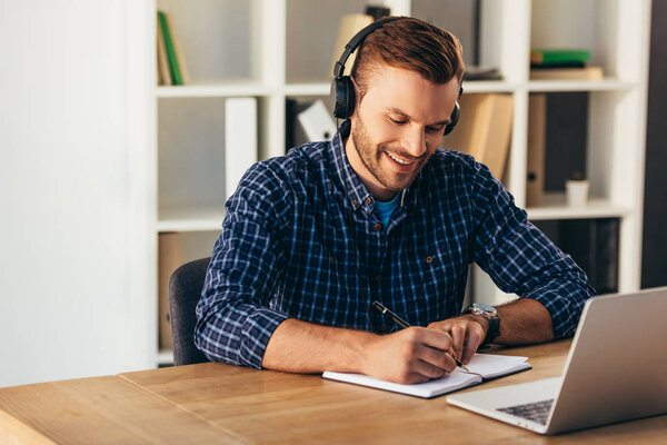 portrait of smiling man in headphones making notes while taking part in webinar at tabletop with laptop in office