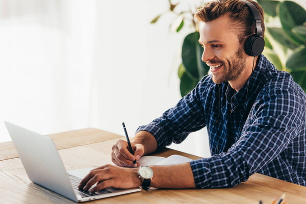 улыбающийся человек в наушниках принимает участие в вебинаре за столом с ноутбуком в офисе
