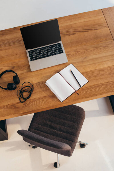 top view of laptop with blank screen, headphones and notebook on wooden table with office chair near by
