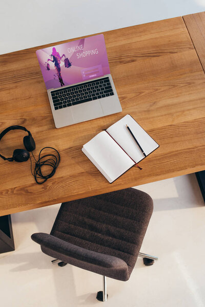 top view of laptop with online shopping lettering on screen, headphones and notebook on wooden table with office chair near by