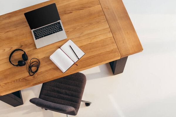 top view of laptop with blank screen, headphones and notebook on wooden table with office chair near by