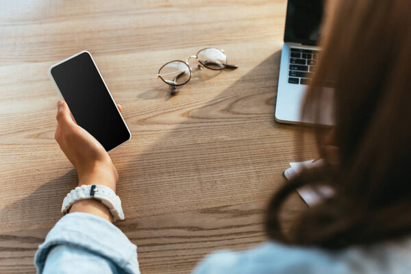 частичный вид женщины со смартфоном, участвующей в вебинаре за столом с ноутбуком
