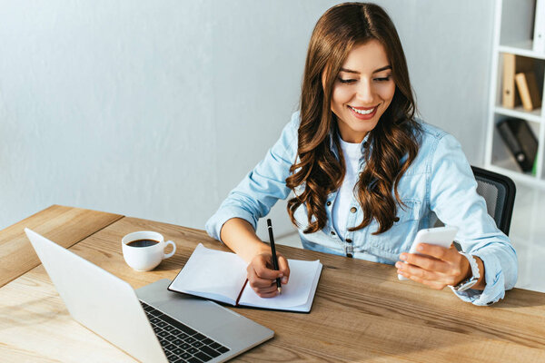 улыбающаяся женщина со смартфоном за столом с ноутбуком принимает участие в вебинаре в офисе
