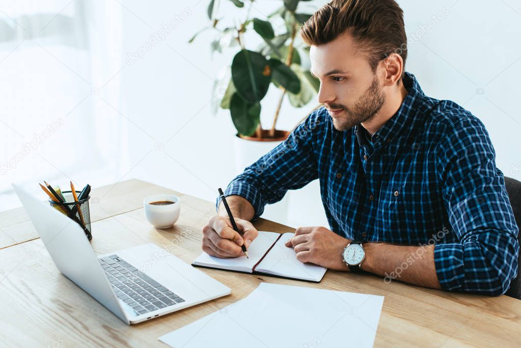 businessman making notes while taking part in webinar at tabletop with laptop in office