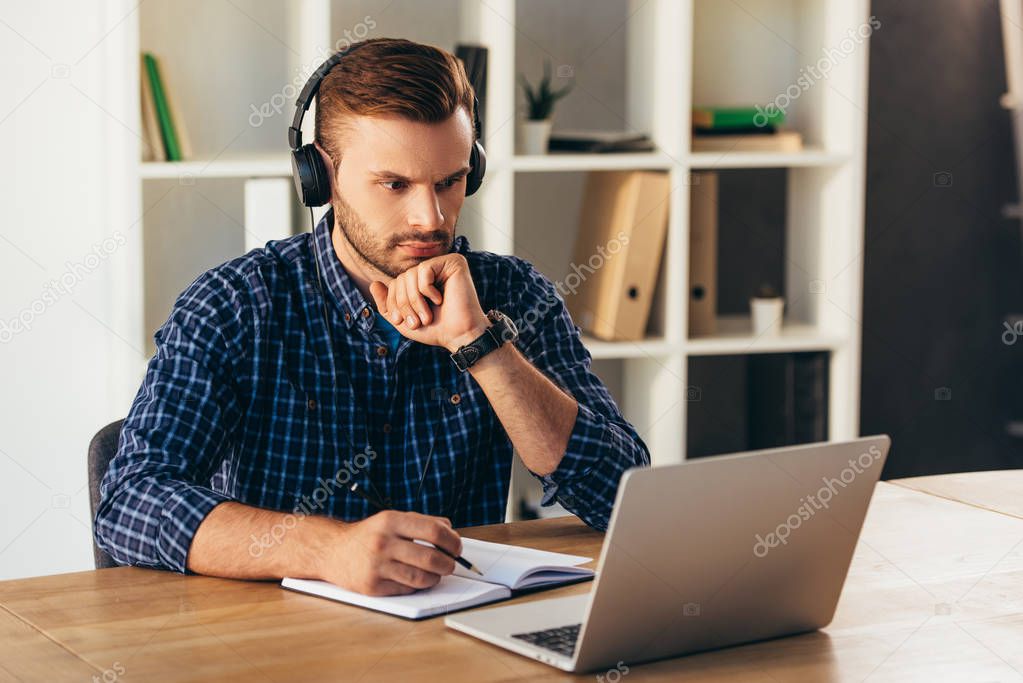 portrait of concentrated man in headphones taking part in webinar at tabletop with notebook in office