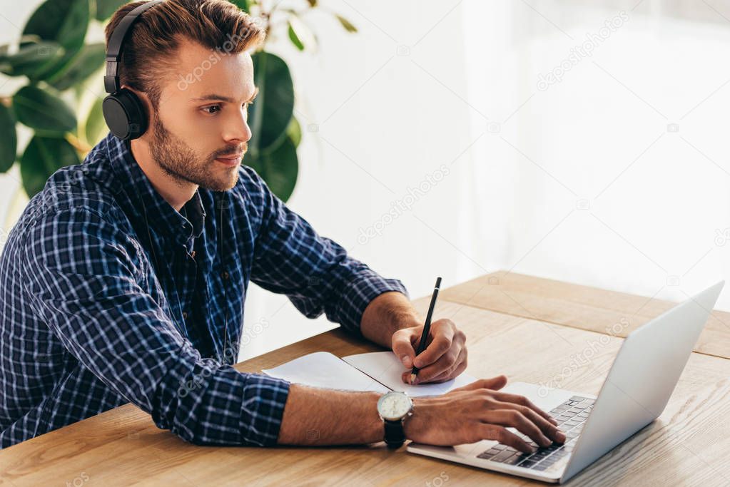 side view of man in headphones taking part in webinar at tabletop with notebook in office