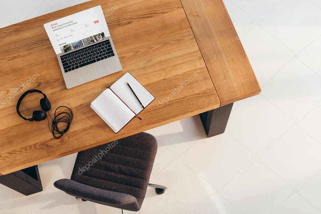 Top view of laptop with airbnb website, headphones and notebook on wooden table with office chair near by