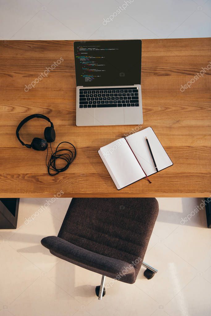 Top view of laptop, headphones and notebook on wooden table with office chair near by