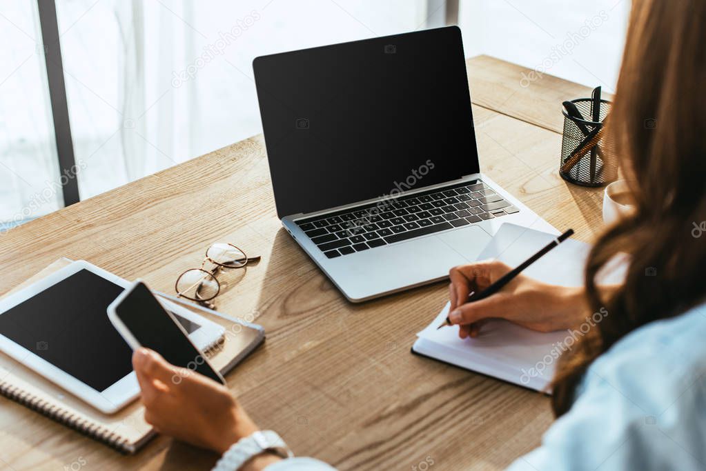 partial view of woman with smartphone taking part in webinar at tabletop with laptop