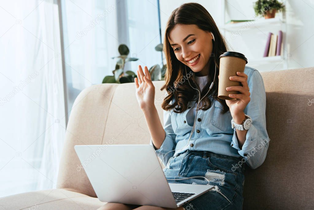 smiling woman in earphones with coffee to go on sofa taking part in webinar at home