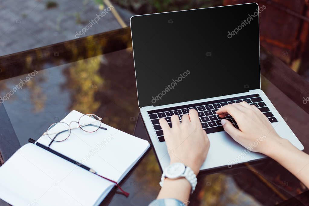 cropped shot of woman working on laptop with blank screen at tabletop with notebook and eyeglasses while taking part in webinar