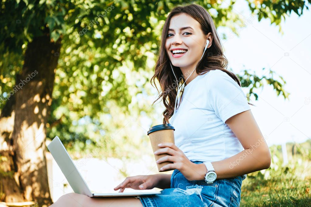 smiling beautiful girl taking part in webinar with laptop in park, looking at camera