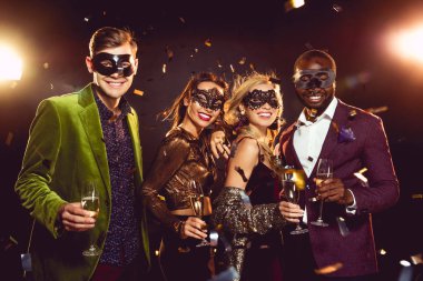 glamorous multiethnic friends in carnival masks holding champagne glasses and celebrating new year on party clipart