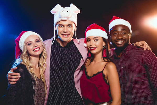 smiling man in pig hat posing with multicultural friends in santa hats on black with backlit 