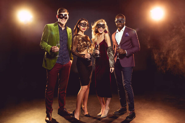 гламурные мультикультурные друзья в карнавальных масках, держащие бокалы для шампанского на вечеринке с задним светом
