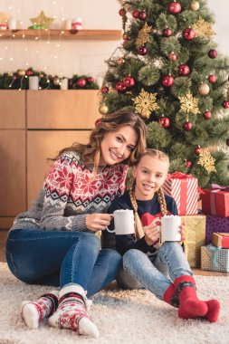 Anne ve kızı evde kapuçino Noel ağacı yakınındaki bardak tutan gülümseyerek